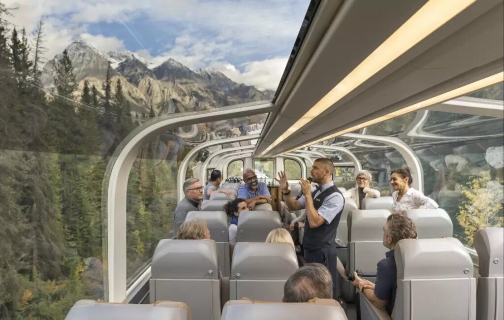 Inside view of The Rocky Mountaineer train in Canada, featuring a man speaking into a microphone while passengers listen. The glass-roofed train offers stunning views of snow-capped mountains outside