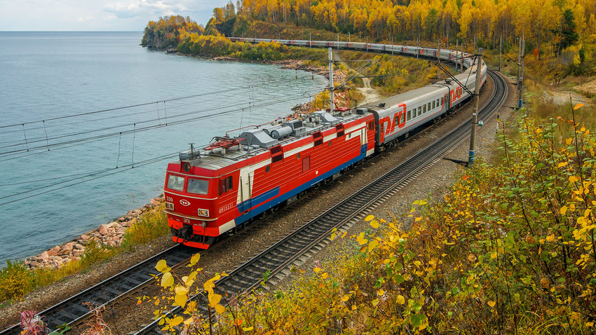 The Trans-Siberian Railway train running alongside flowing water, with a dense forest of tall trees visible on the opposite side