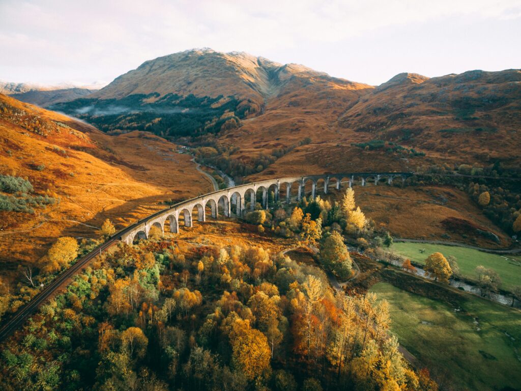 Aerial view of The West Highland Line in Scotland, showcasing a train track on a bridge surrounded by towering trees and mountains on the left, with lush green land on the right