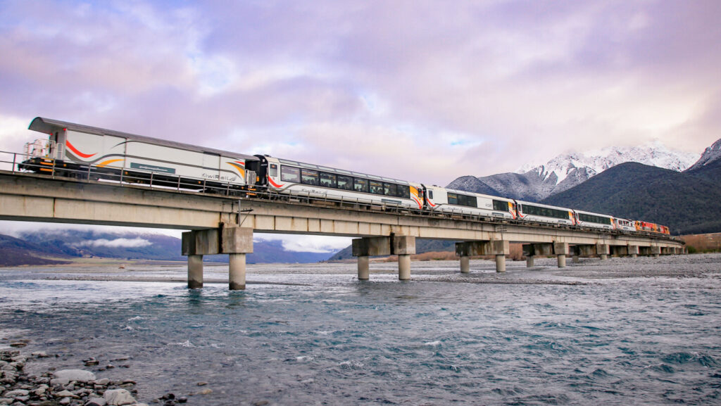 The TranzAlpine train crossing a bridge in New Zealand, with flowing water below and snow-capped mountains visible in the background, partially shrouded in fog